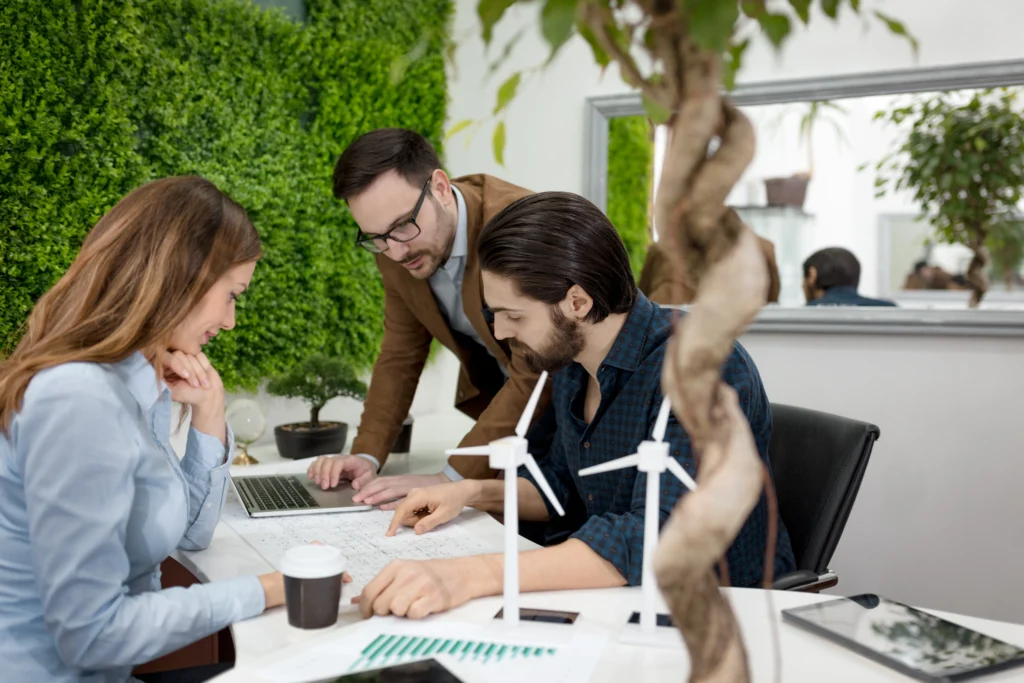 Un equipo colabora en un espacio lleno de vegetación natural, con turbinas eólicas sobre la mesa, un entorno donde se puede aplicar perfectamente qué es un sistema de gestión ambiental en la práctica.