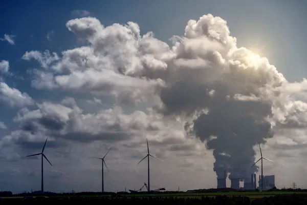 Cielo cubierto por grandes columnas de humo junto a molinos de viento, mostrando el contraste entre energías limpias y las emisiones que intensifican el efecto invernadero.