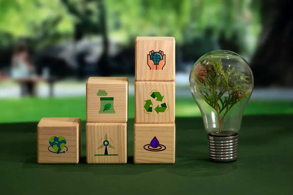 Bloques de madera con íconos de reciclaje, energía y sostenibilidad junto a un foco con una planta verde, representando cómo las empresas integran el valor compartido.